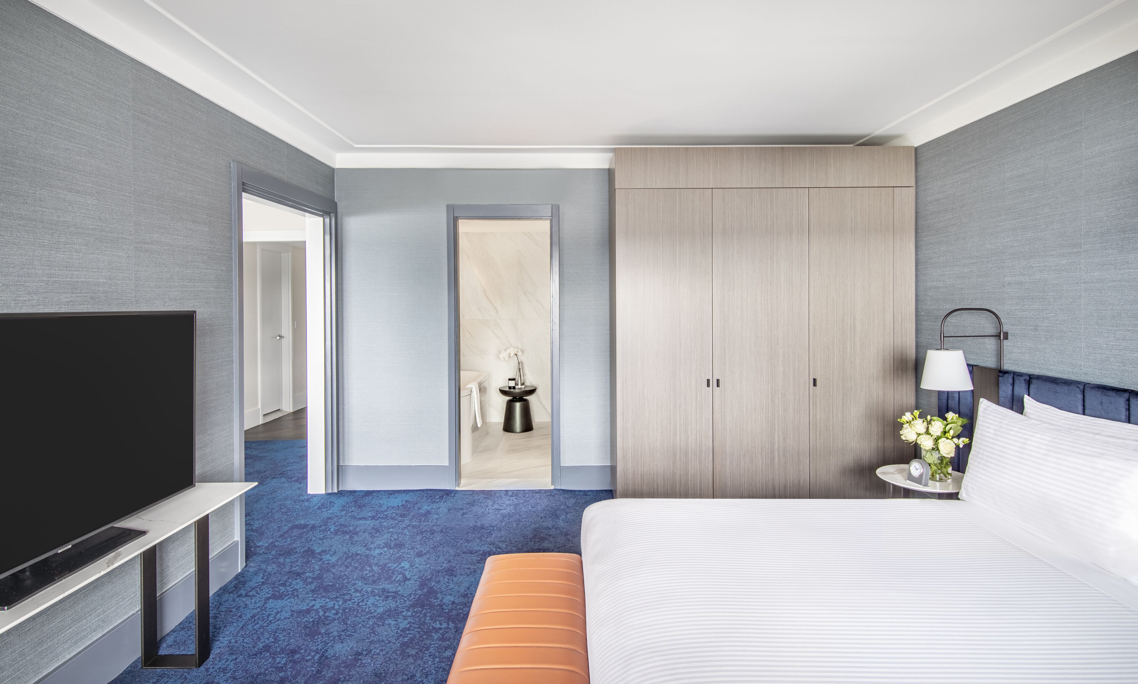 A modern hotel room with a large flat-screen TV, a bed with white linens, a blue carpet, a wooden wardrobe, and a small bedside table with a lamp and flowers.