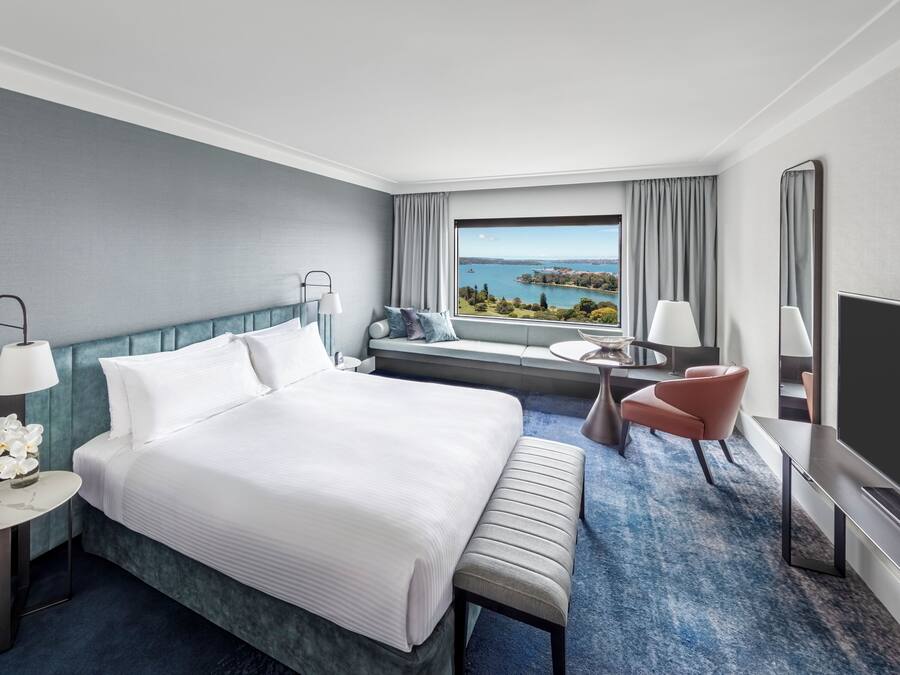 Habitación de hotel con una cama grande, zona de estar con vistas, televisor y una mesita de noche con lámpara.