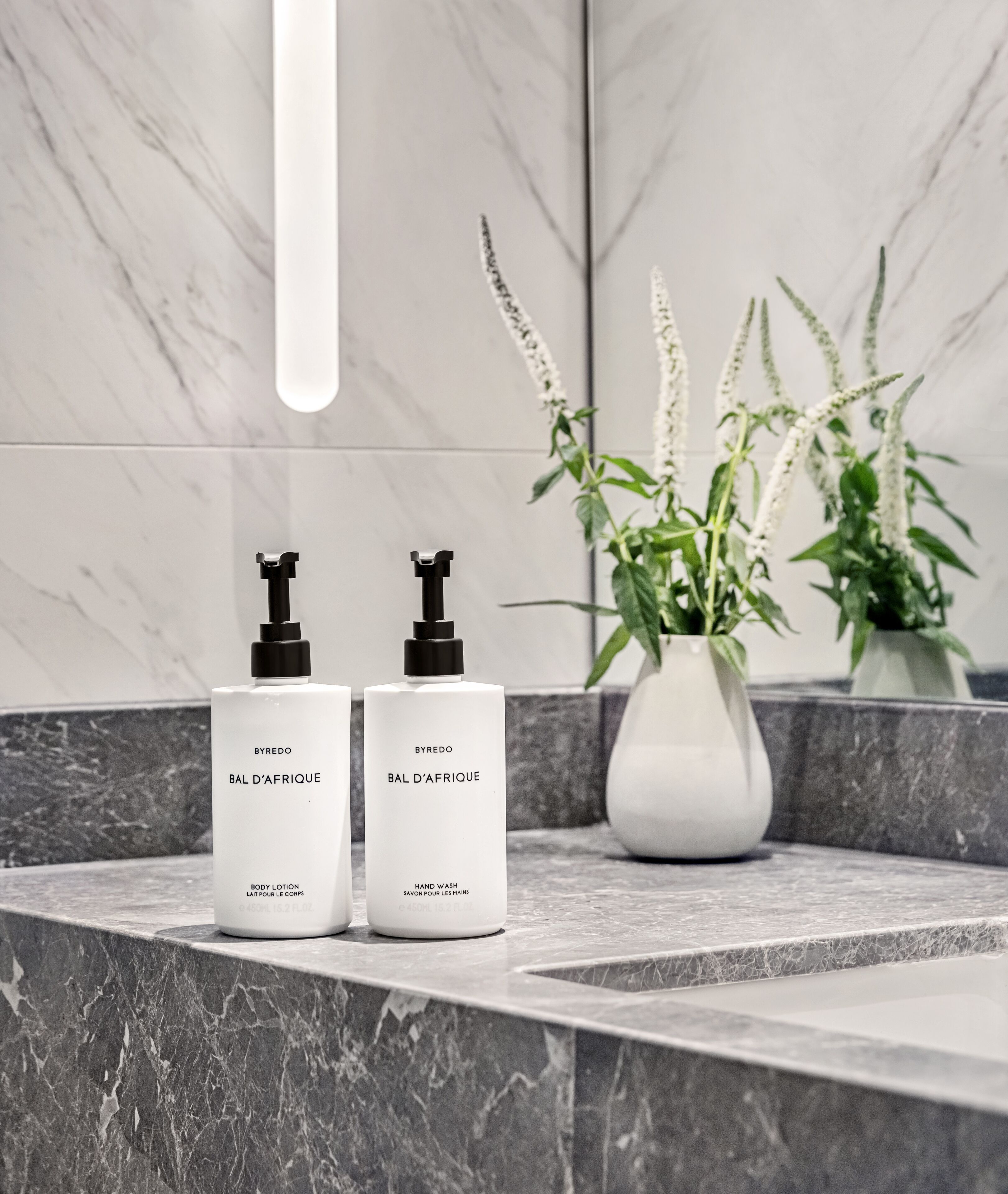 Two white BYREDO BAL D'AFRIQUE body lotion and hand wash bottles on a marble countertop with a white vase containing green plants.