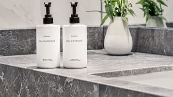 Two white BYREDO BAL D'AFRIQUE body lotion and hand wash bottles on a marble countertop with a white vase containing green plants.