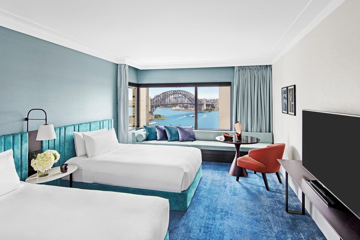 A hotel room with two beds, a TV, a small table, and a view of a bridge.