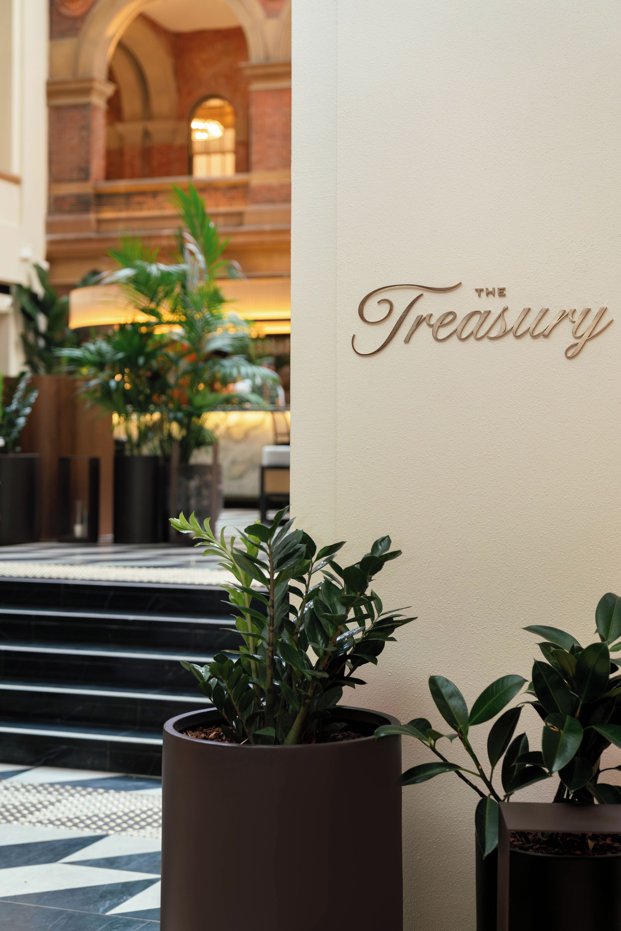 A lobby with a large potted plant, a wall with the inscription 'THE Treasury', and a staircase leading to an upper level.