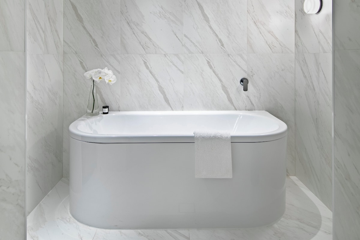 A modern bathroom with a freestanding bathtub, marble walls, and a small vase with flowers.