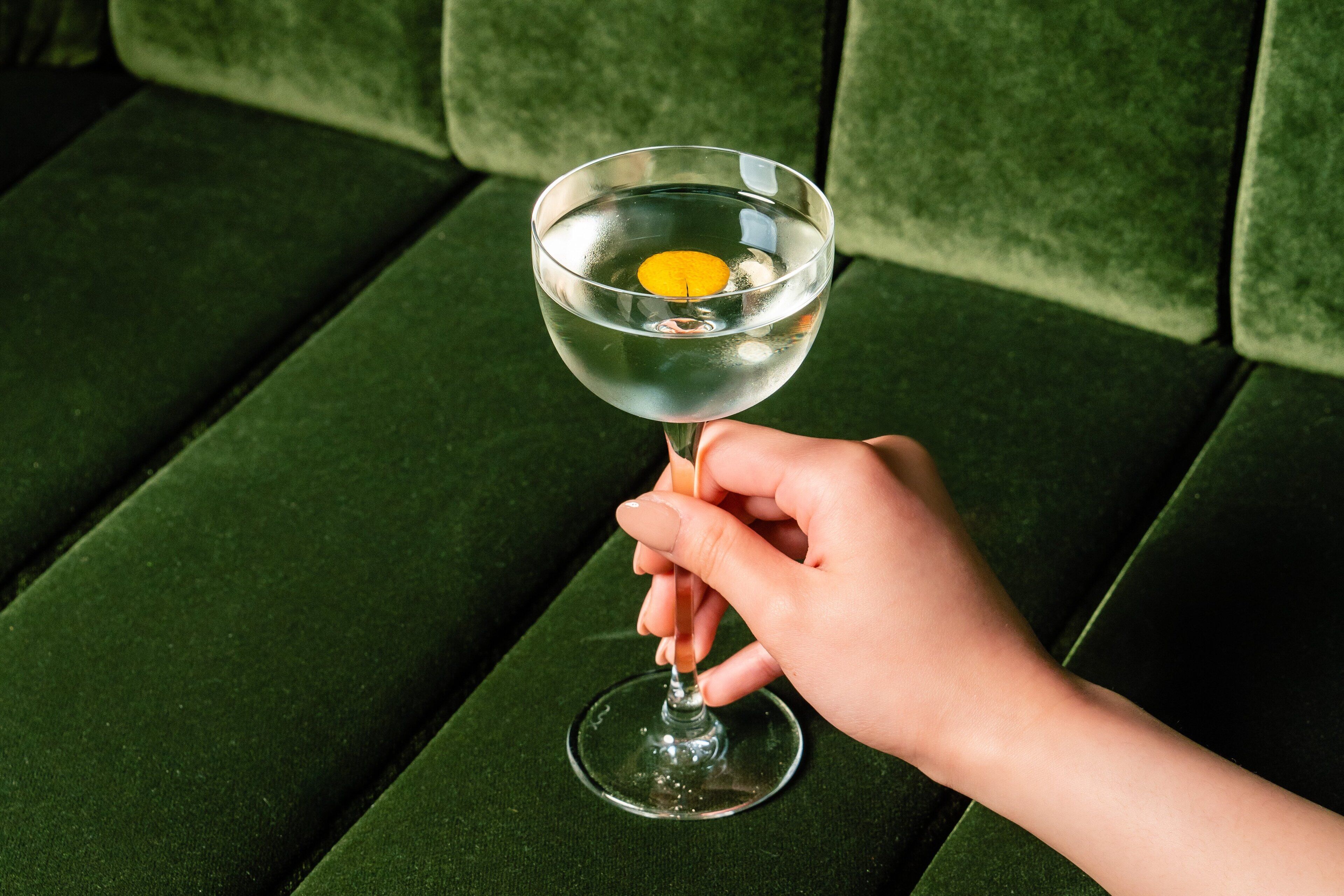 A hand holding a glass with a lemon slice, placed on a green upholstered surface.