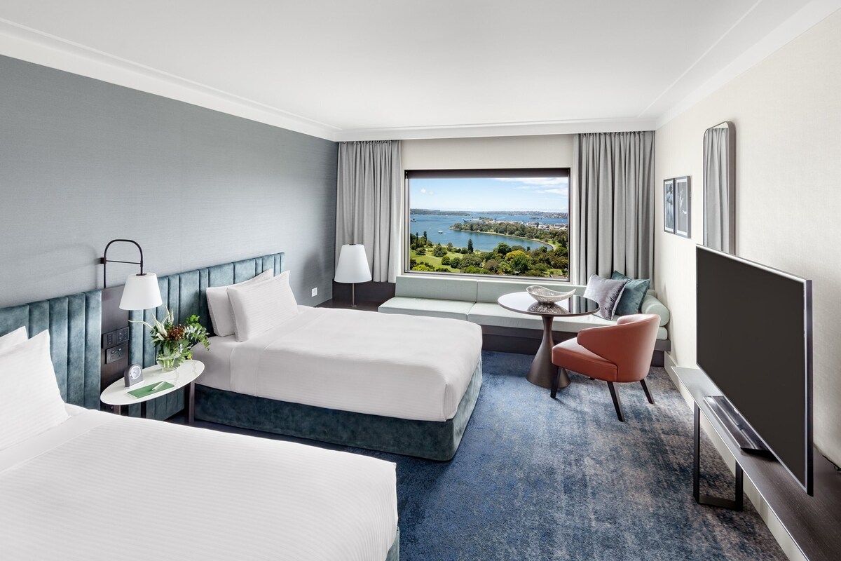 A hotel room with two beds, a TV, a seating area with a table, and a view of a body of water.