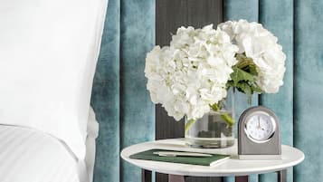 Una cama bien hecha con ropa de cama blanca, una mesita de noche con una lámpara blanca, un jarrón con flores blancas y un reloj.