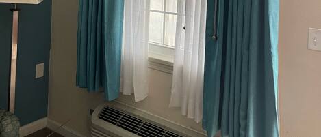 A room with a window covered by teal and white curtains, a standing lamp, and a wall-mounted air conditioner.