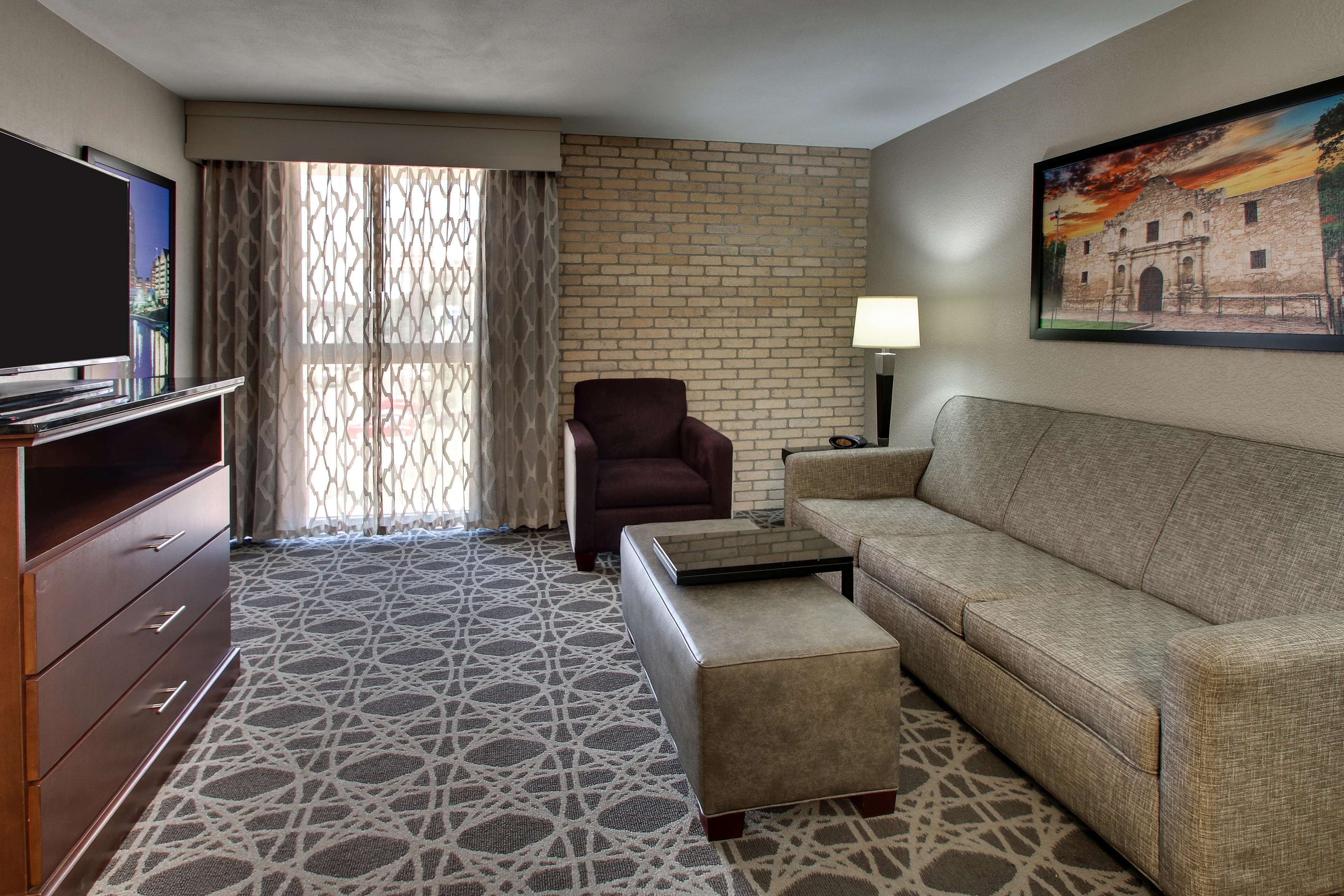A hotel room with a brick wall, a large window with curtains, a flat-screen TV, a sofa, a chair, a coffee table, and a painting of a building.