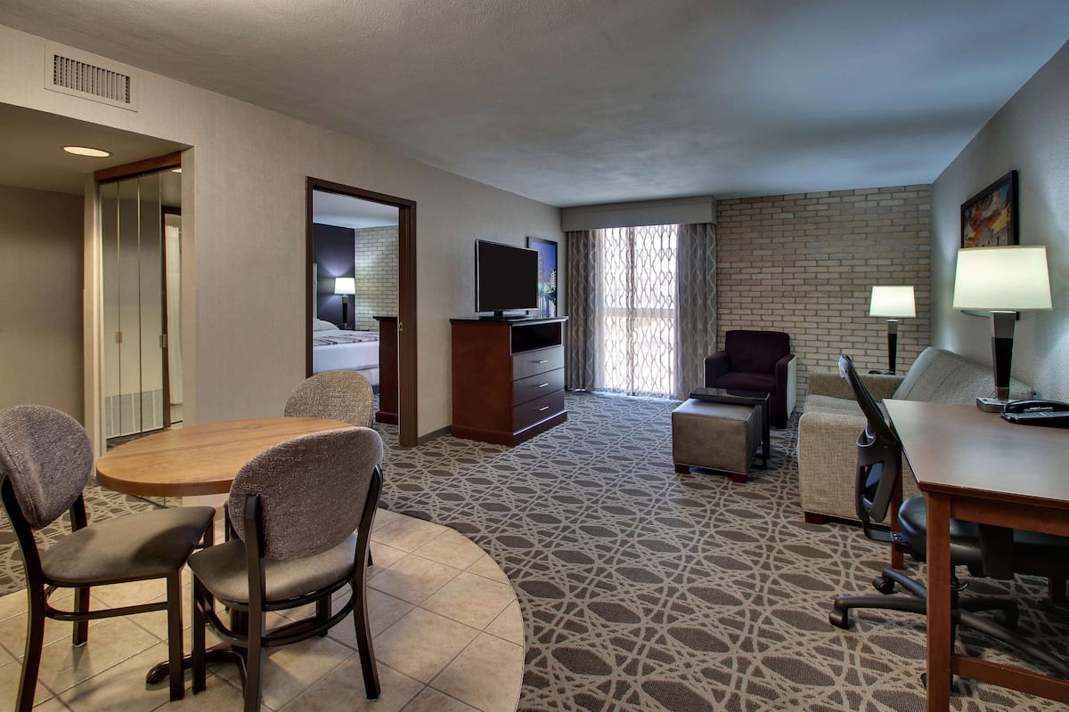 A hotel room with a dining table, chairs, a TV, a desk with a lamp, and a patterned carpet.