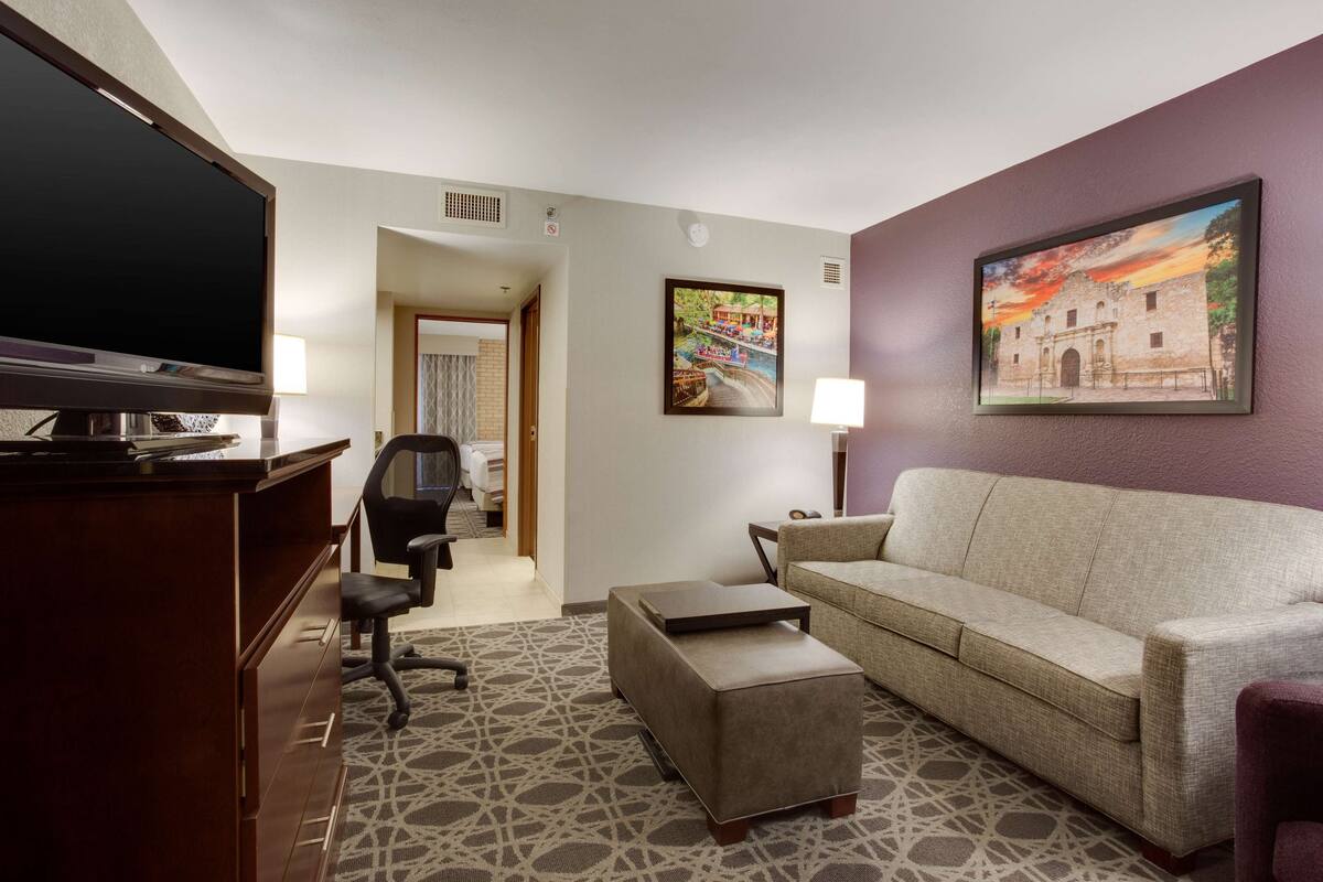 A hotel room with a large flat-screen TV, a sofa, a coffee table, and a patterned rug.
