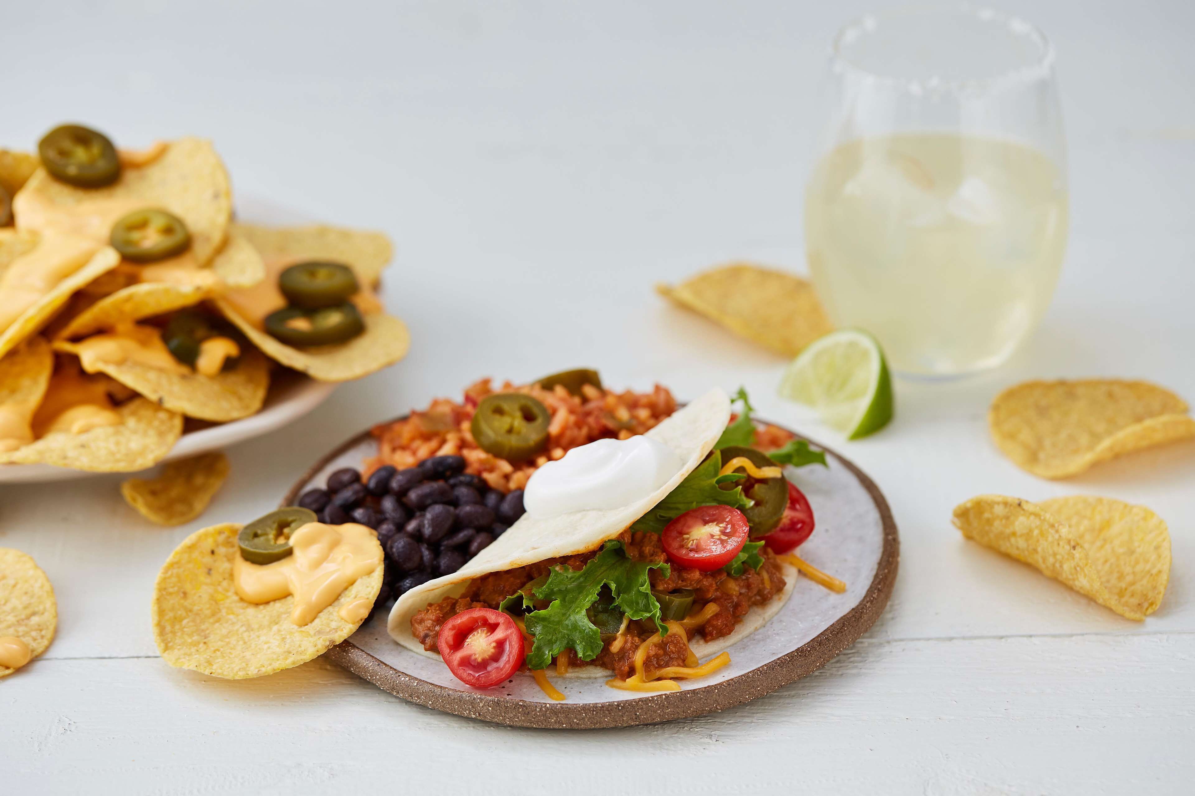 A plate of nachos, a taco with toppings, and a glass of white beverage on a white surface.