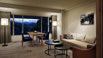 A hotel room with a dining area, a sofa, a coffee table, and a view of mountains through large windows.