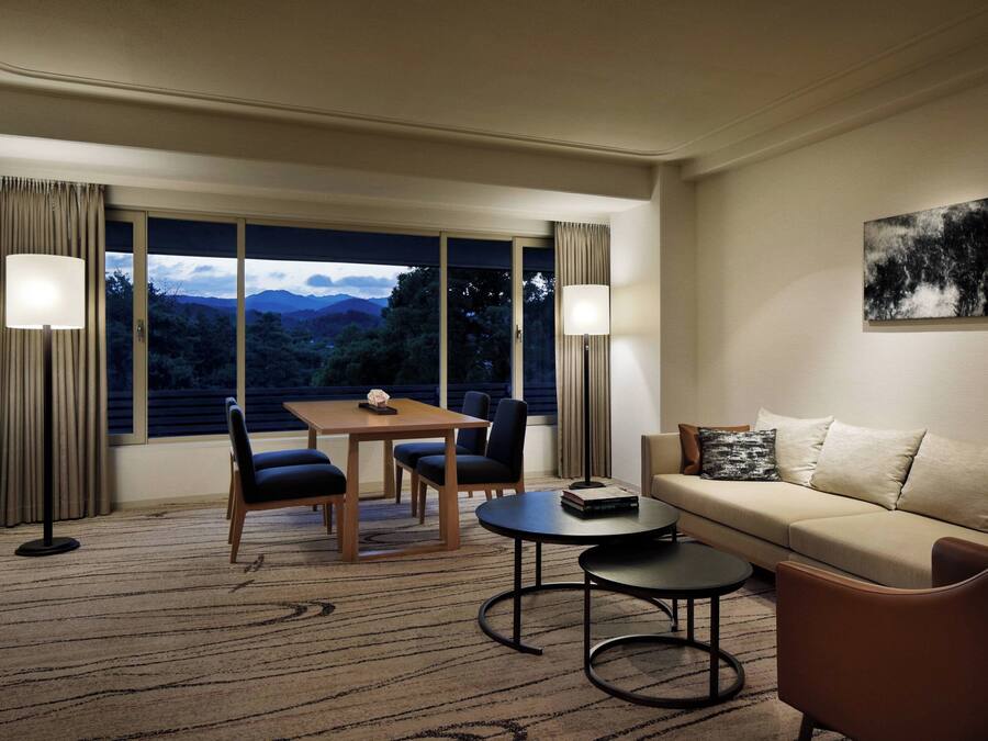 A hotel room with a dining area, a sofa, a coffee table, and a view of mountains through large windows.
