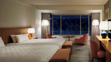 A hotel room with two beds, a desk, a chair, a small table, and a view of the outdoors through a large window.