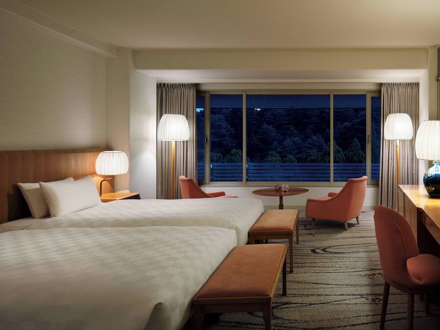 A hotel room with two beds, a desk, a chair, a small table, and a view of the outdoors through a large window.