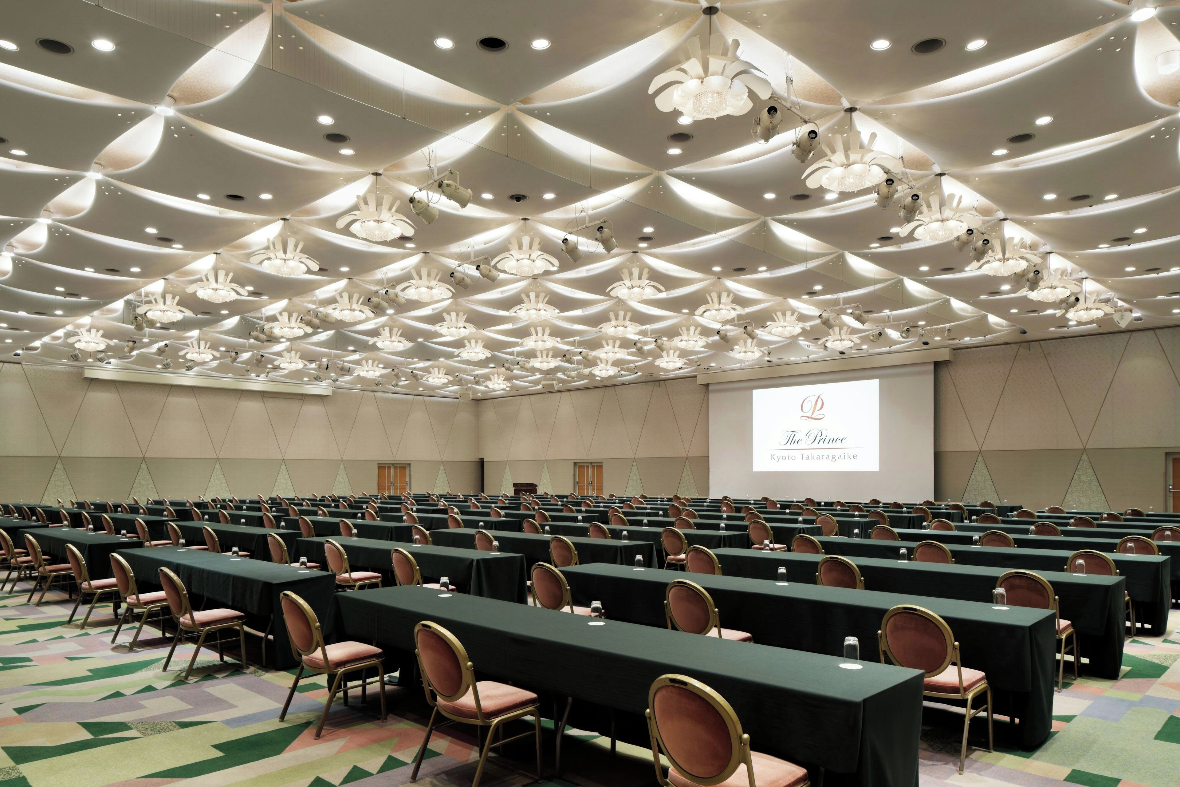 A conference room with rows of tables and chairs, a large screen, and a ceiling with unique lighting fixtures.