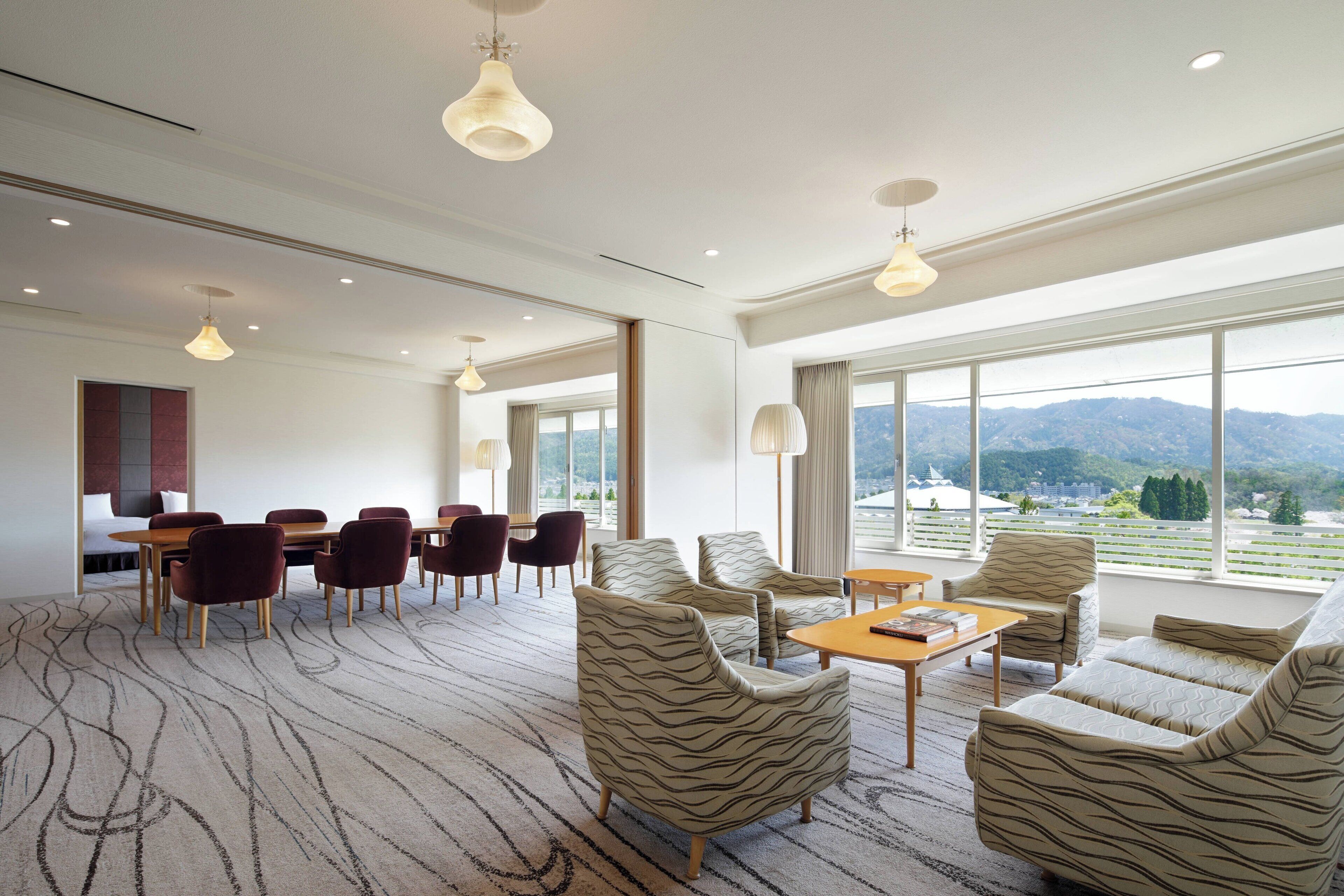 A spacious hotel room with a large window offering a mountain view, a dining area with a table and chairs, and patterned armchairs.