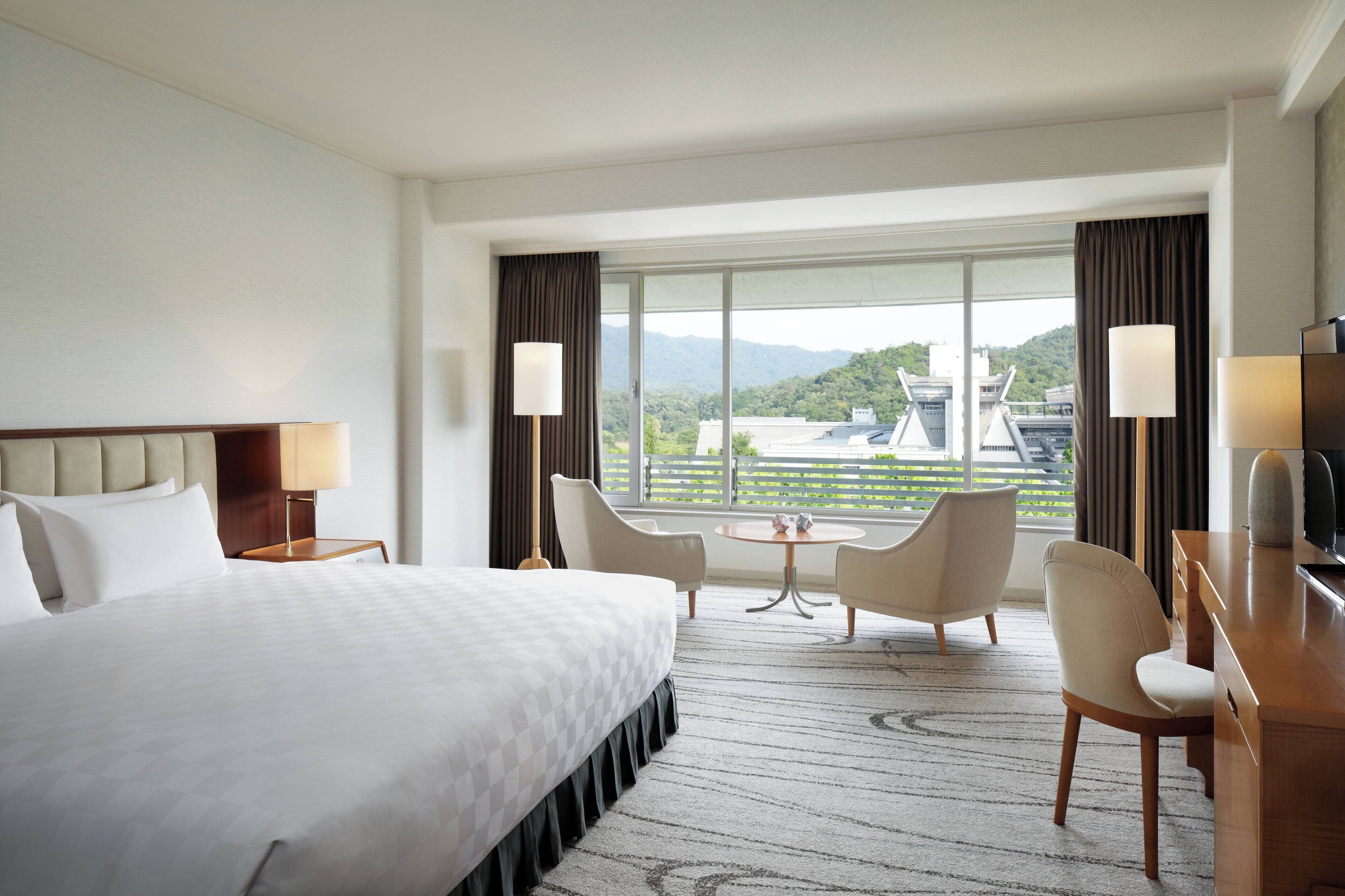 A hotel room with a large bed, two chairs, a small table, and a desk. There is a window with a view of buildings and greenery.