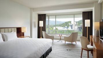 A hotel room with a large bed, two chairs, a small table, and a desk. There is a window with a view of buildings and greenery.