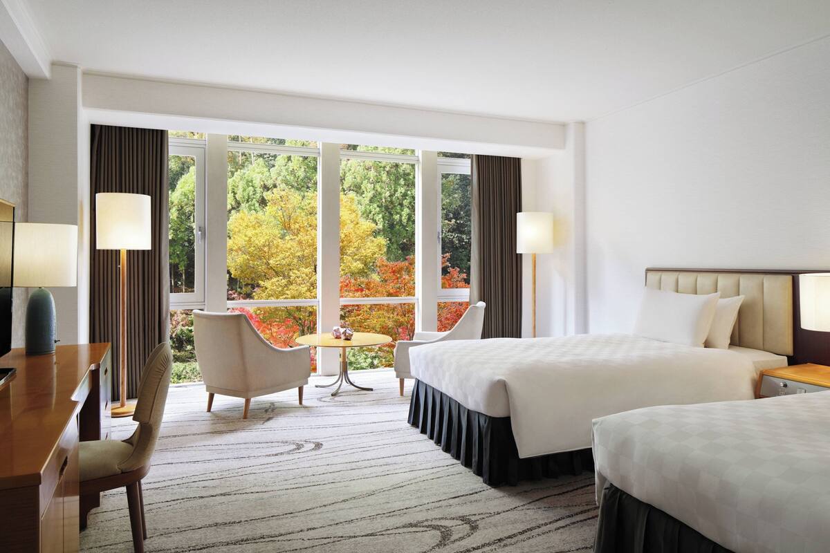 Ein Hotelzimmer mit zwei Betten, einem Schreibtisch, einem Stuhl, einem kleinen Tisch und großen Fenstern mit Blick auf Herbstlaub.