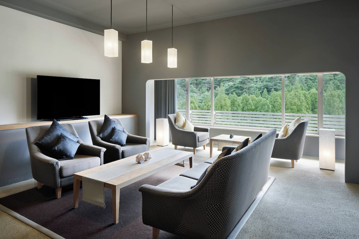 Ein modernes Wohnzimmer mit einem Flachbildfernseher, grauen Sofas mit schwarzen Kissen, einem Holztisch und einem großen Fenster mit Blick auf Bäume.