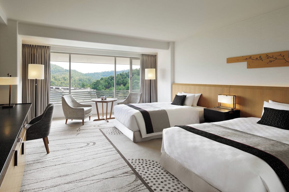 Ein Hotelzimmer mit zwei Betten, einem Schreibtisch, einem Sessel, einem kleinen Tisch und Blick auf eine bewaldete Landschaft.