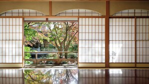 Ein traditioneller japanischer Raum mit Tatami-Bodenbelag, Schiebetüren und Blick auf einen Garten mit Bäumen und einer Steinbrücke.