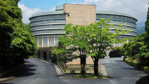 Ein großes, rundes Gebäude mit flachem Dach und zahlreichen Fenstern, umgeben von üppiger Vegetation und einer gepflasterten Straße.