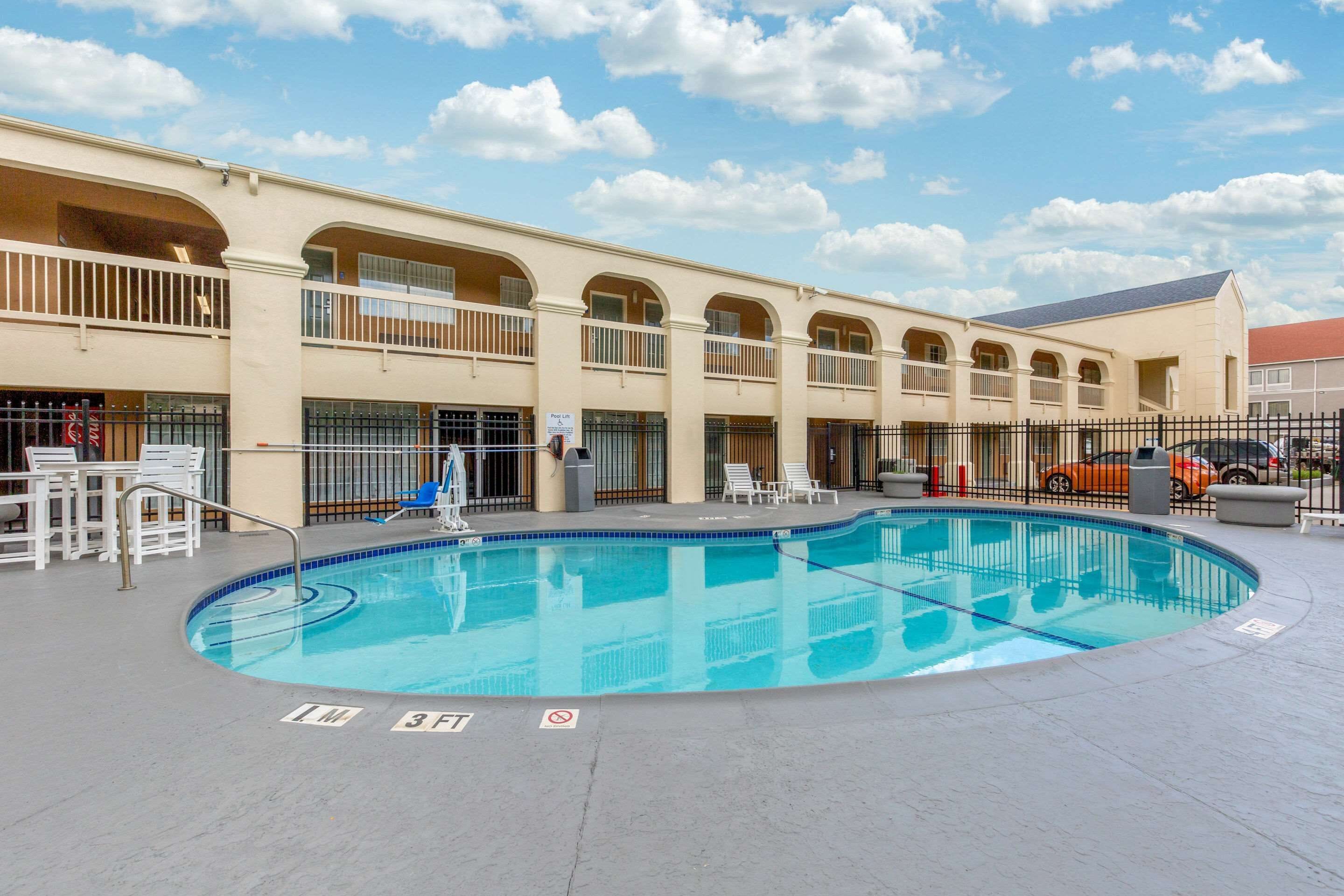 A swimming pool with lounge chairs and a fenced area, surrounded by a building with multiple balconies and a red-roofed structure in the background.