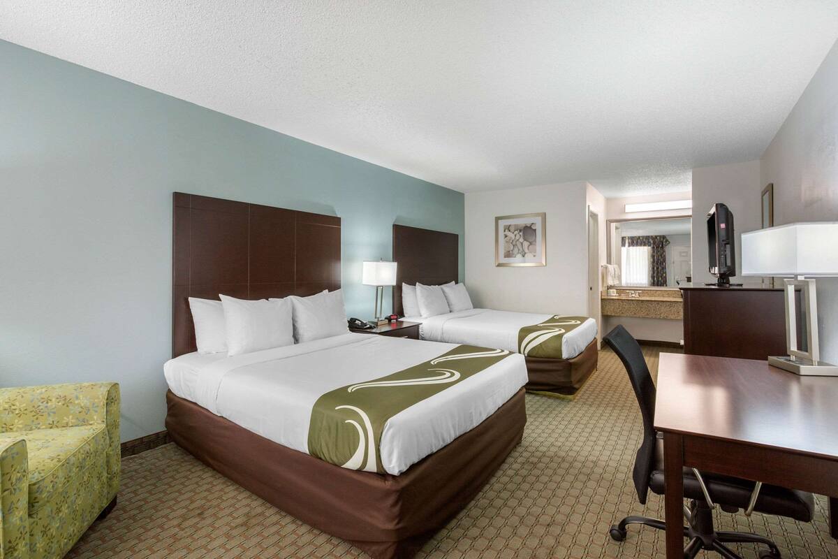 Quarto de hotel com duas camas, uma escrivaninha, uma cadeira, um abajur e um espelho.