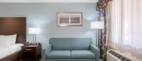 A hotel room with a bed, a sofa, a bedside table with a lamp, a framed picture on the wall, and a window with curtains.