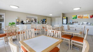 A dining area with multiple tables and chairs, a kitchen in the background, and a refrigerator stocked with beverages. - Quality Inn Clute Freeport (Clute)