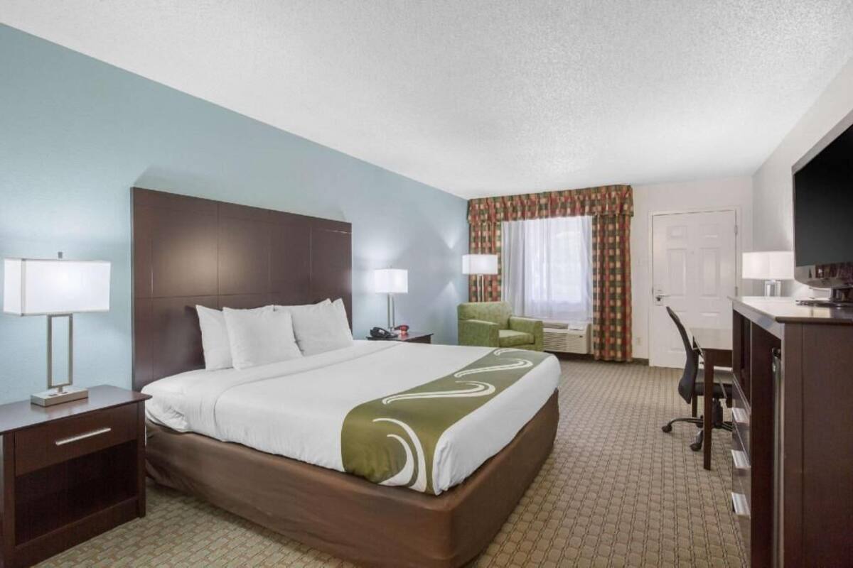 Quarto de hotel com cama, luminárias de cabeceira, escrivaninha, cadeira e televisão.