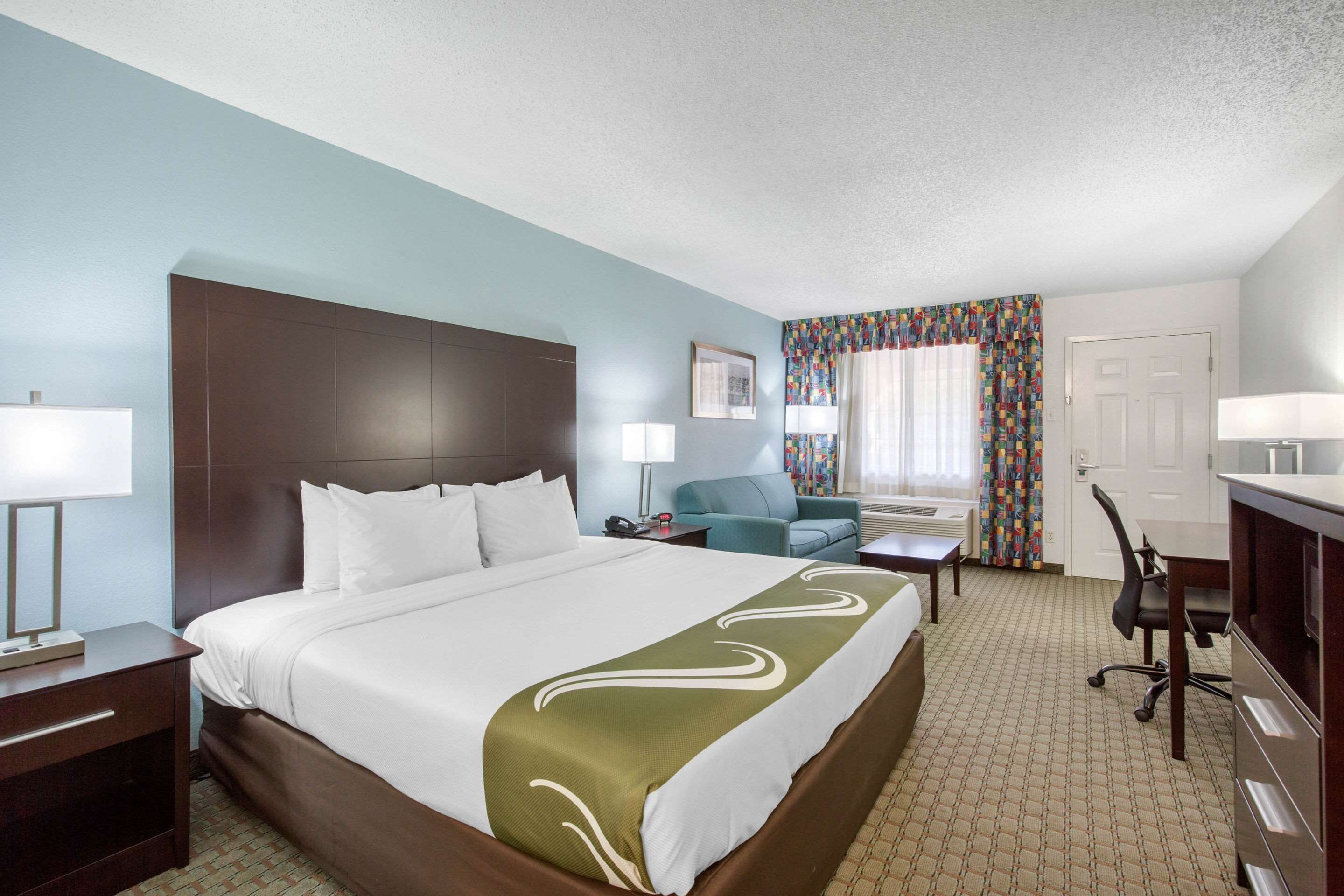 A hotel room with a large bed, a desk, a chair, a couch, and a desk lamp.
