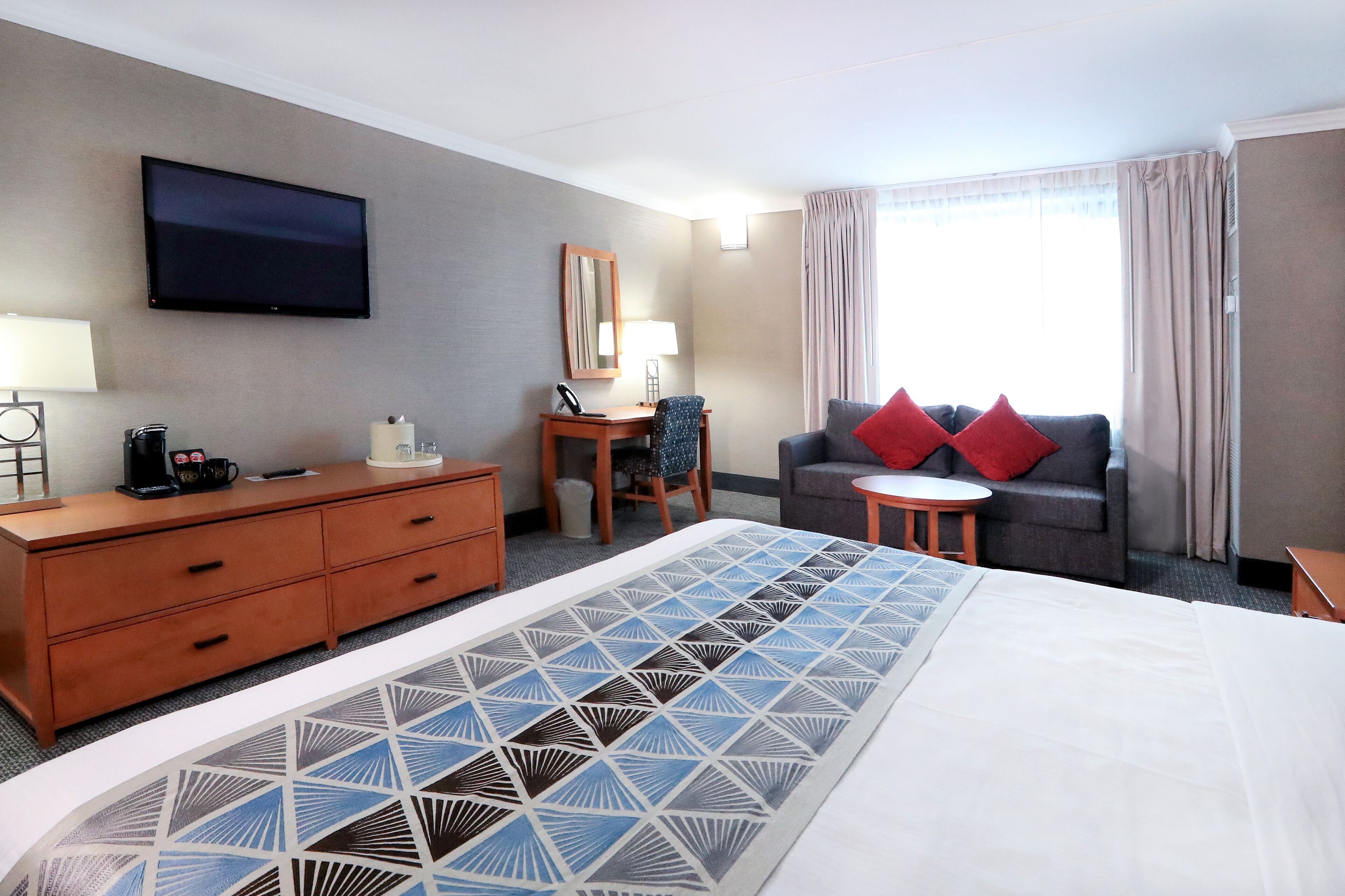 A hotel room with a bed, a desk, a chair, a sofa, a TV, and a window with curtains.