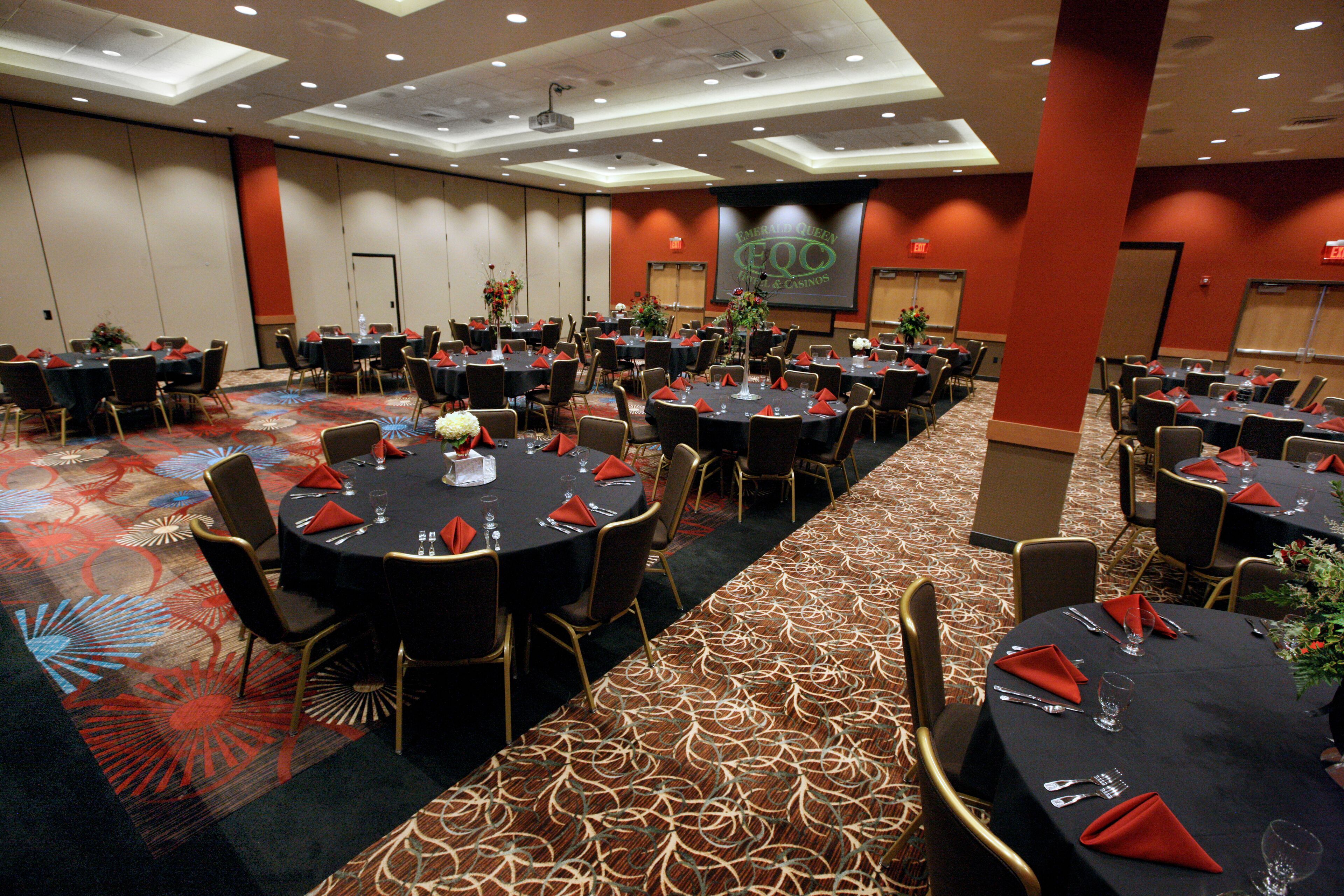A large conference room with multiple round tables, each set with red napkins, glasses, and cutlery.