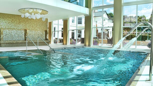 A large indoor swimming pool with clear water, a metal ladder, and a view of a house through large windows. - Bedford Lodge Hotel & Spa (Newmarket)