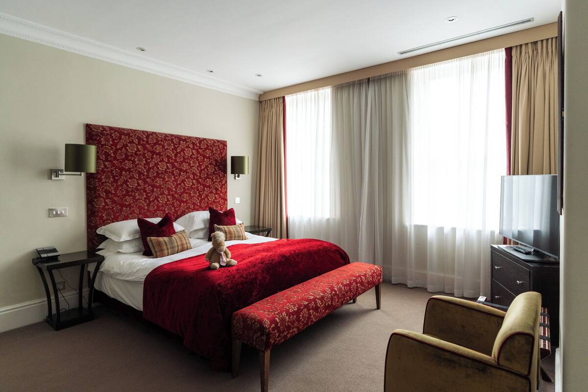Een hotelkamer met een groot bed, een nachtkastje, een stoel, een bankje, een televisie en gordijnen.