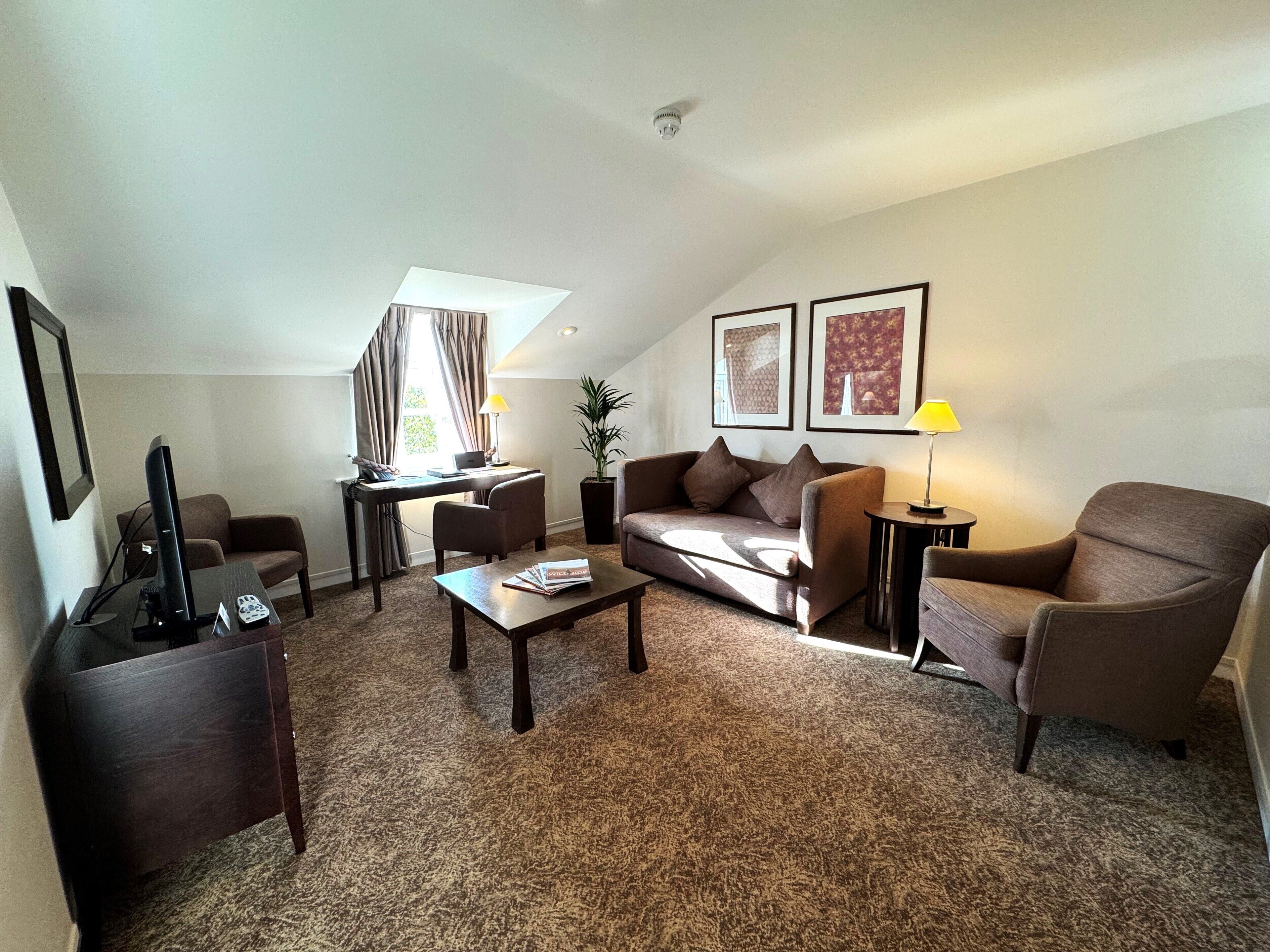 A furnished living room with a sofa, armchairs, a coffee table, a desk with a lamp, and framed pictures on the wall.