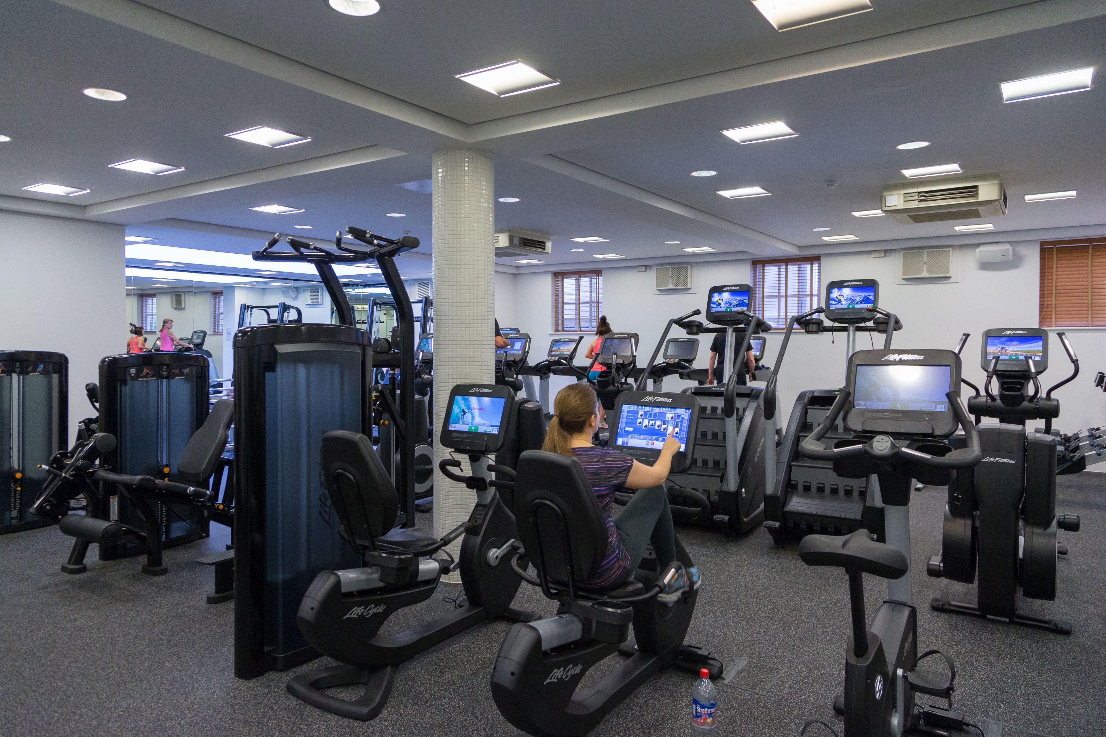 Un gimnasio con diferentes máquinas de ejercicio, incluyendo cintas de correr y elípticas, y una persona usando una bicicleta estática.