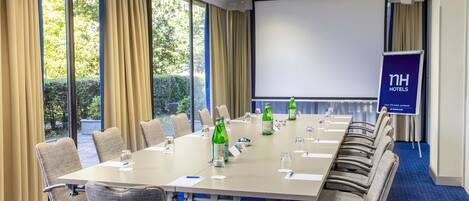 A conference room with a long table, chairs, a projector screen, and a banner for NH Hotels.