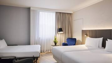 A hotel room with two beds, a desk, a chair, a lamp, and a potted plant.