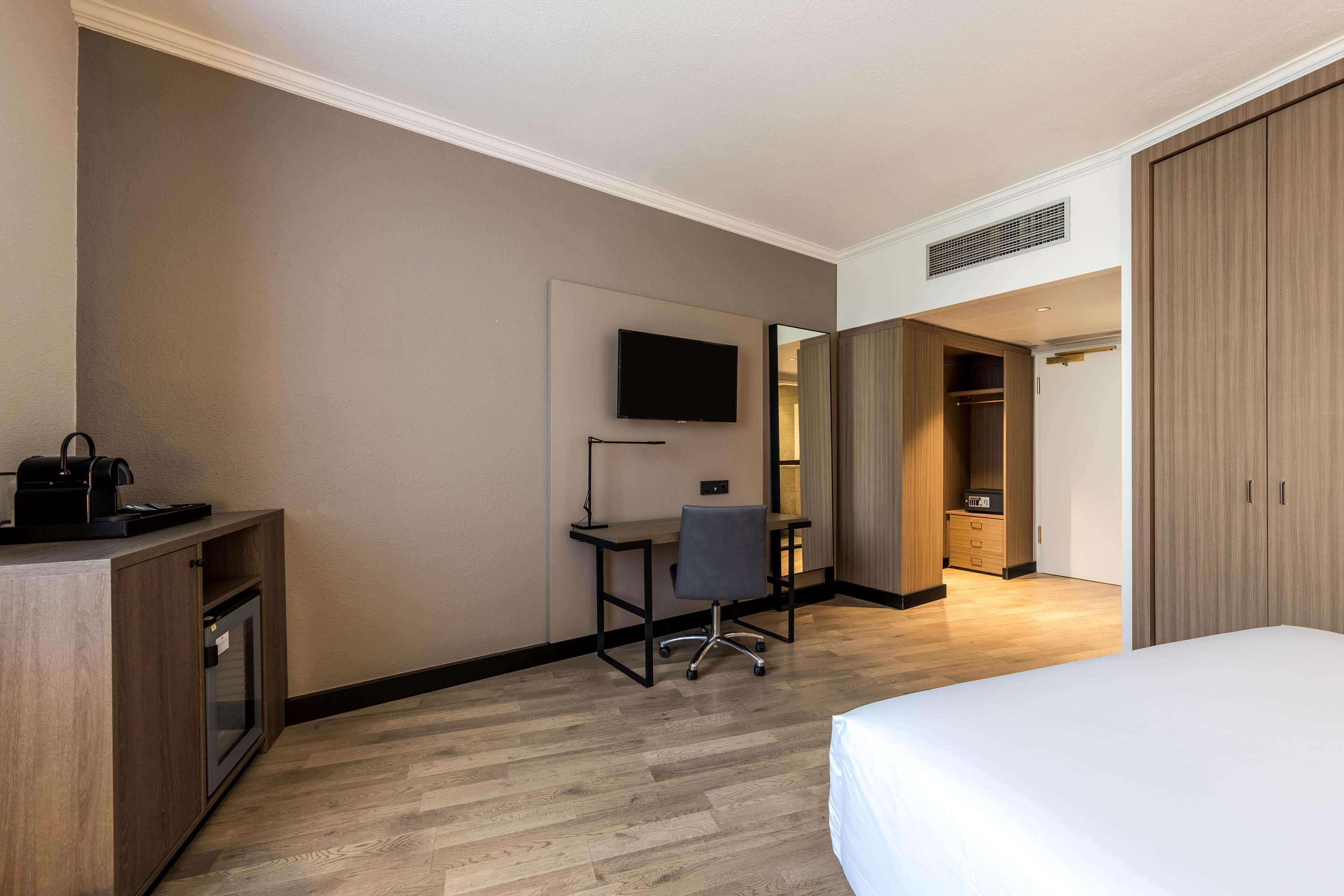 A hotel room with a bed, desk, chair, TV, wardrobe, and a minibar.