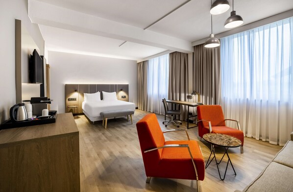 A hotel room with a bed, a desk, two chairs, a table, and a large window with curtains.