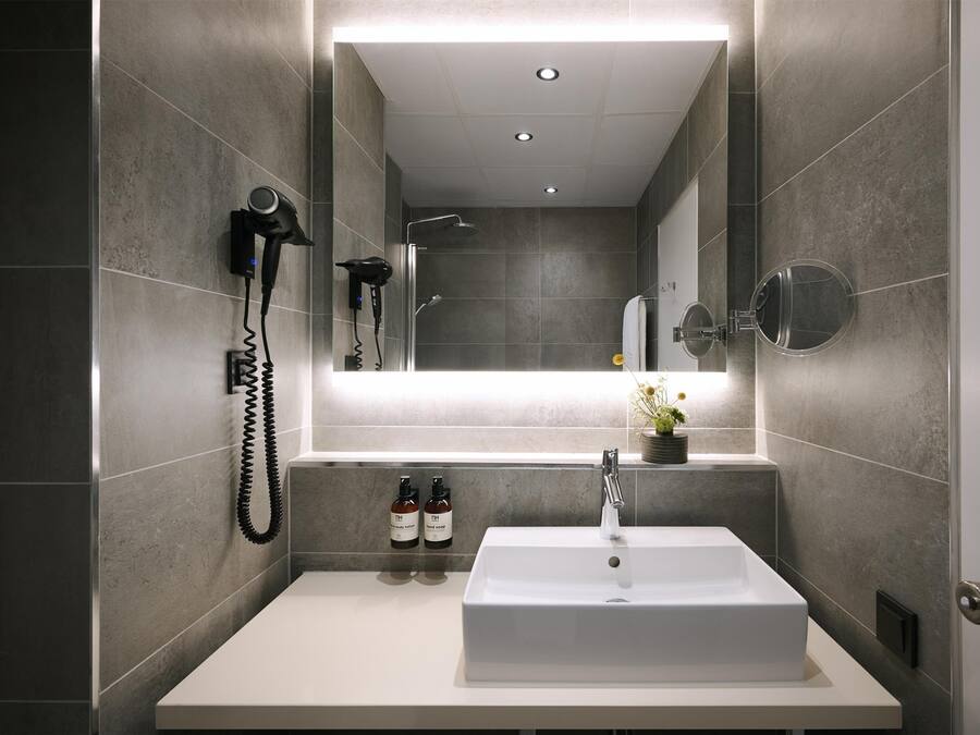 Une salle de bain moderne avec un grand miroir, un lavabo blanc, un sèche-cheveux et deux bouteilles de shampoing.