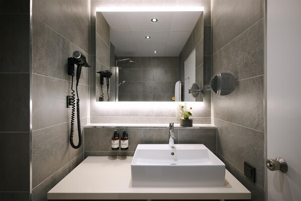 A modern bathroom with a large mirror, a white sink, a handheld hair dryer, and two bottles of shampoo.