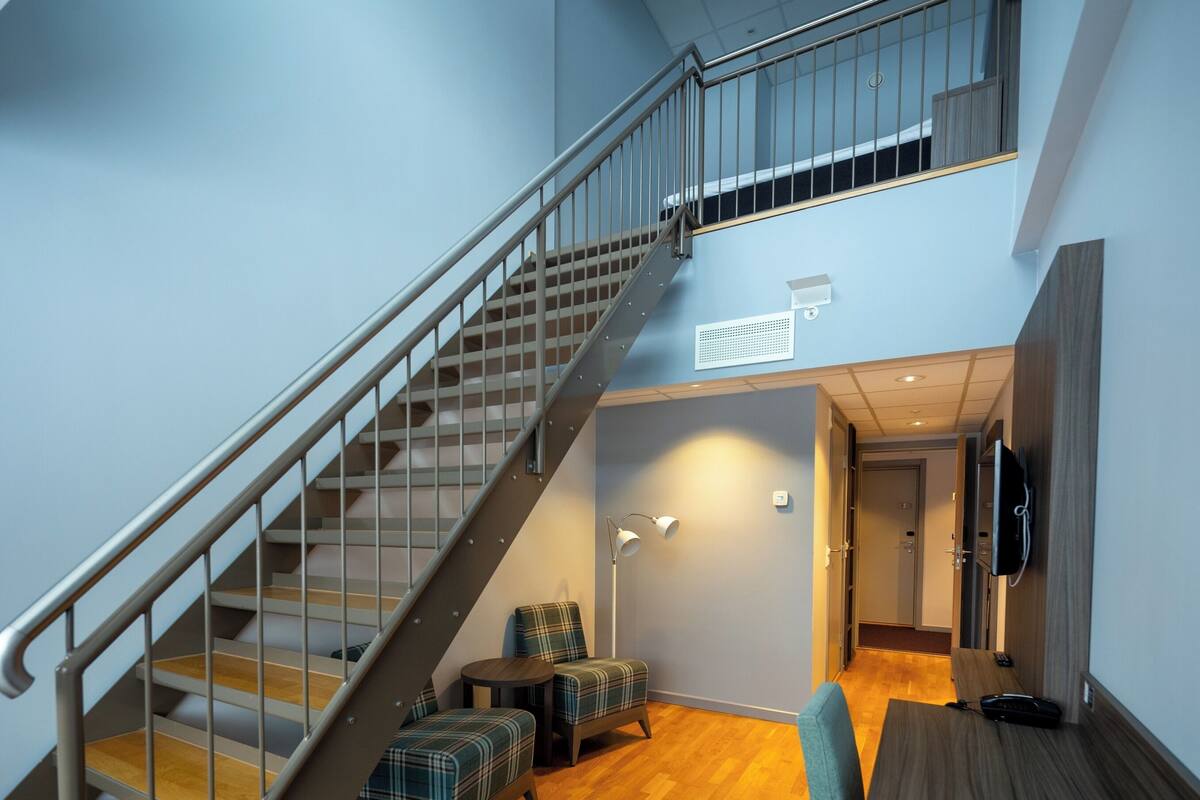Eine Treppe mit Metallgeländern führt zu einer oberen Etage, ein Sitzbereich mit Tisch und Stühlen, ein an der Wand befestigtes Flachbildfernsehen und ein Holzboden.