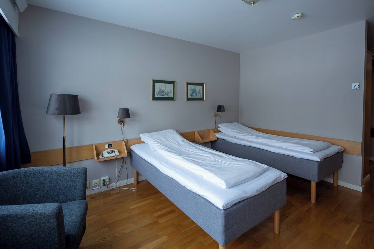 Ein Hotelzimmer mit zwei Einzelbetten, einem Sessel, einem Nachttisch mit Lampe sowie zwei gerahmten Bildern an der Wand.