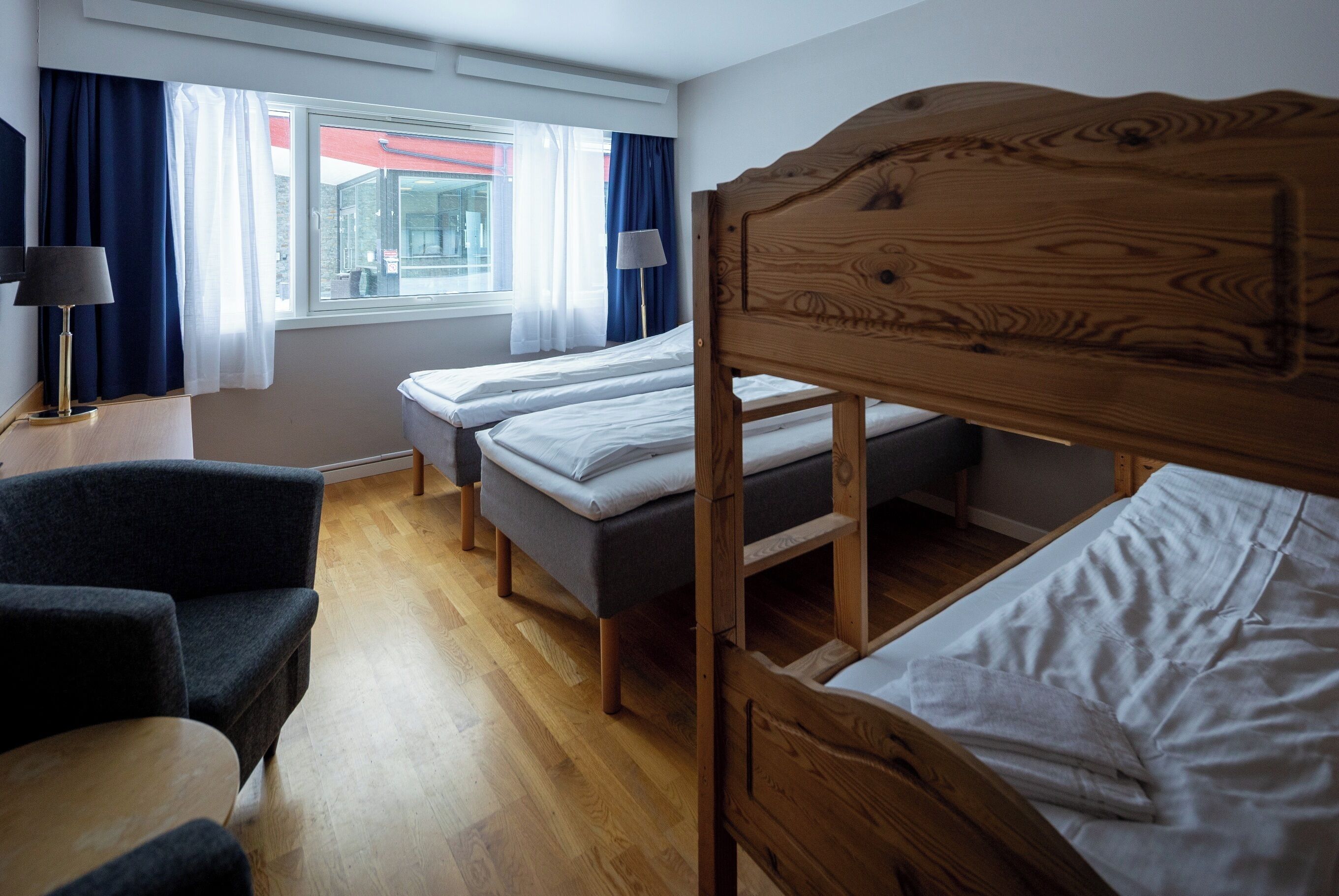 Et hotelværelse med en køjeseng i træ, to enkelt senge, en sofa, et lille bord og et vindue med gardiner.