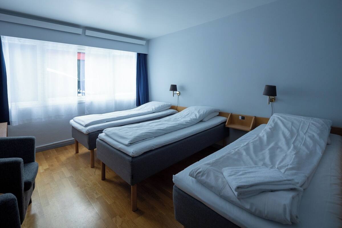 Ein Hotelzimmer mit zwei Einzelbetten, einem Schlafsofa, Holzboden, einem Fenster mit Vorhängen und zwei Wandlampen.